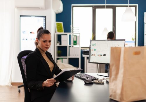 Businesswoman in corporate start up office analyziing documents, chart looking at clipboard. Recycled paper bag with delicious tasty food for lunch break. Employee holding coffee cup.