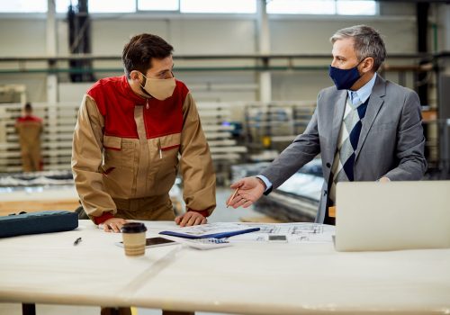 Mid adult businessman and male worker discussing about project plans while wearing protective face masks at carpentry workshop.