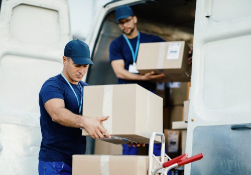 Young courier and his colleague unloading cardboard boxes from delivery van.